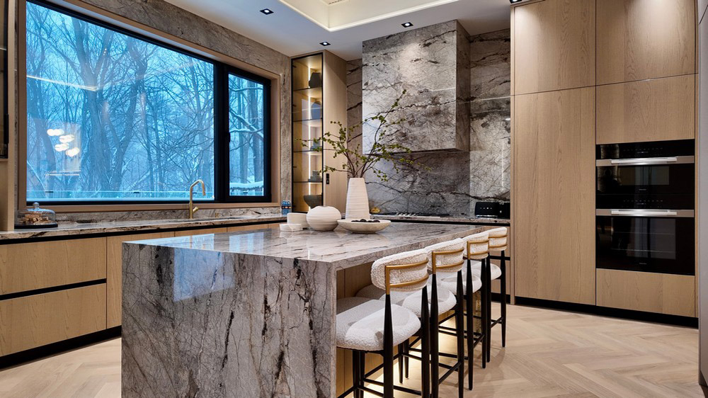 Marble island with seating framed by a picture window and Miele built-in ovens